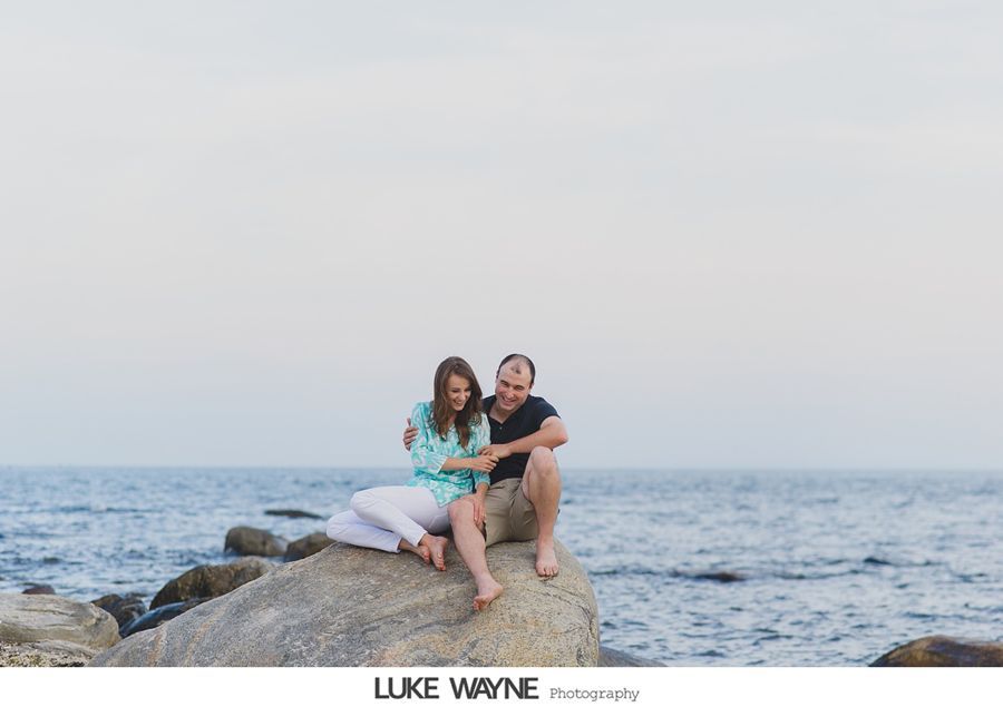 Couple sitting on a large rock by the ocean; woman in turquoise, man in black shirt.