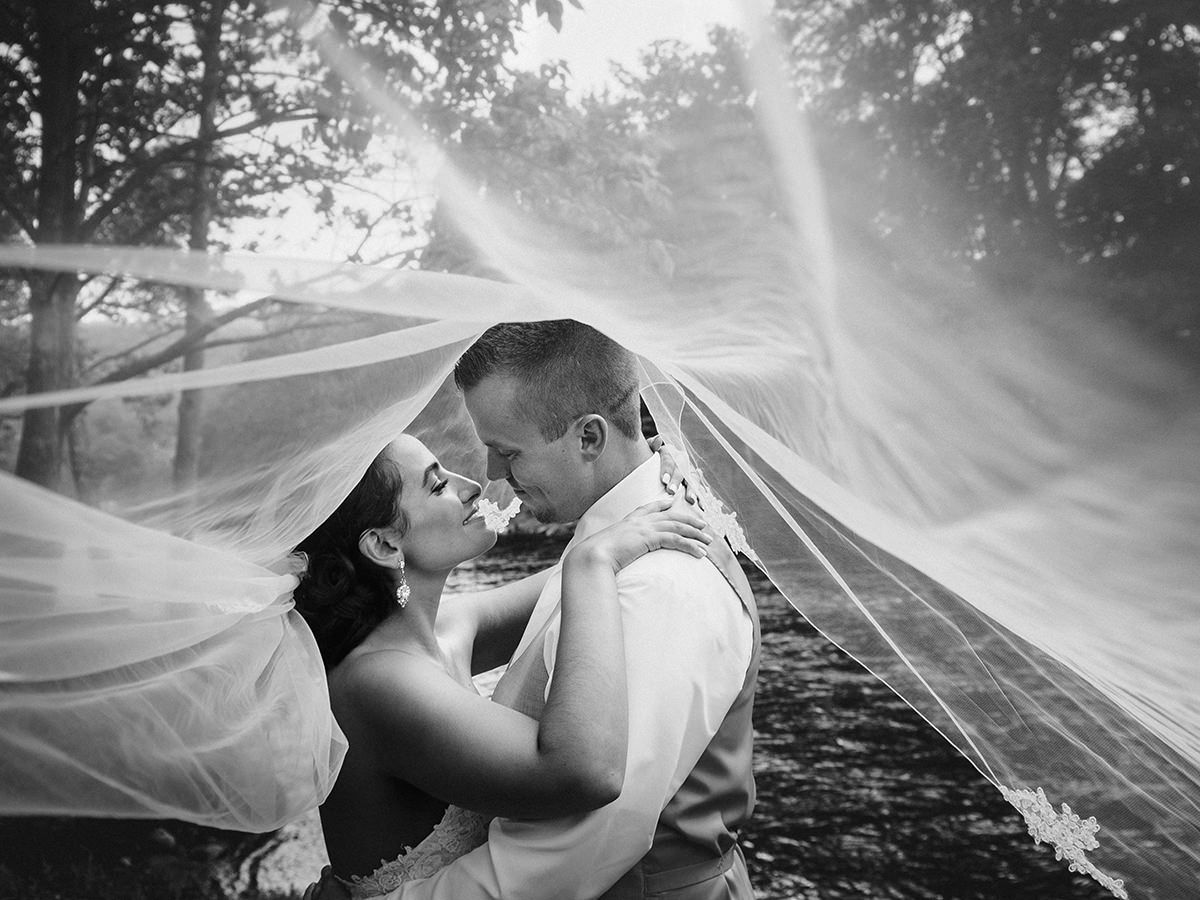 Couple kissing under flowing veil; outdoor setting with trees in black and white.