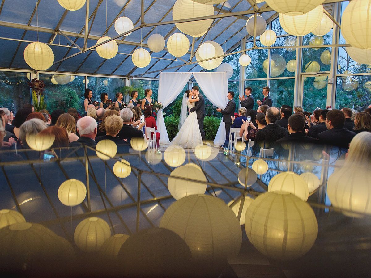 Wedding ceremony in a greenhouse with the couple under a white draped altar; round paper lanterns overhead.