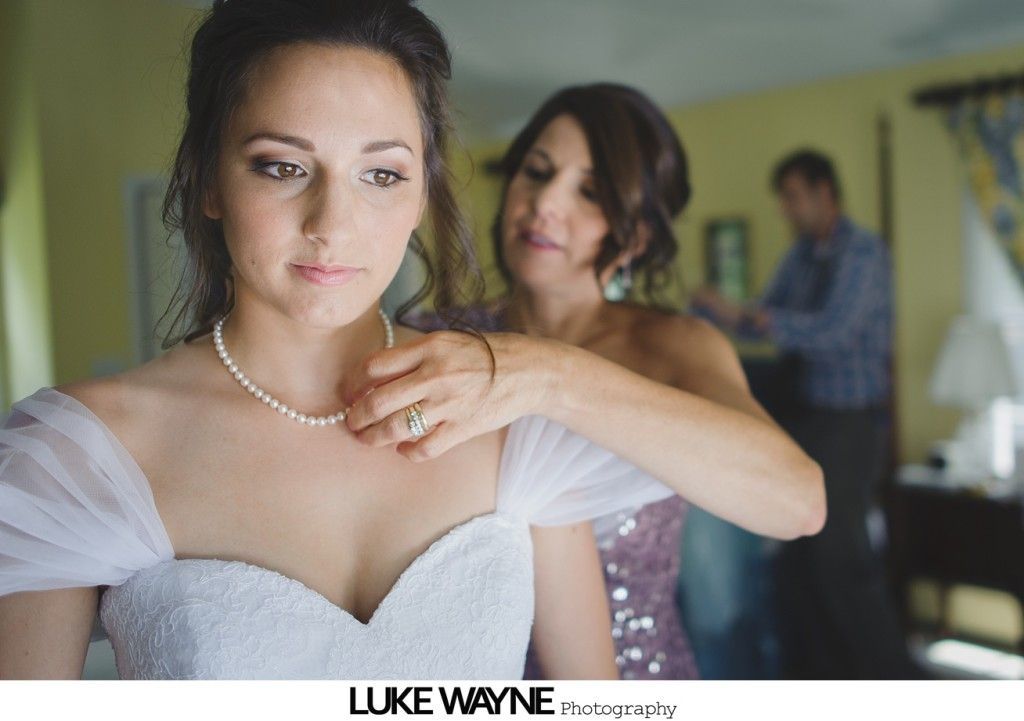 Bride getting her pearl necklace fastened by a woman in a room, man in the background.
