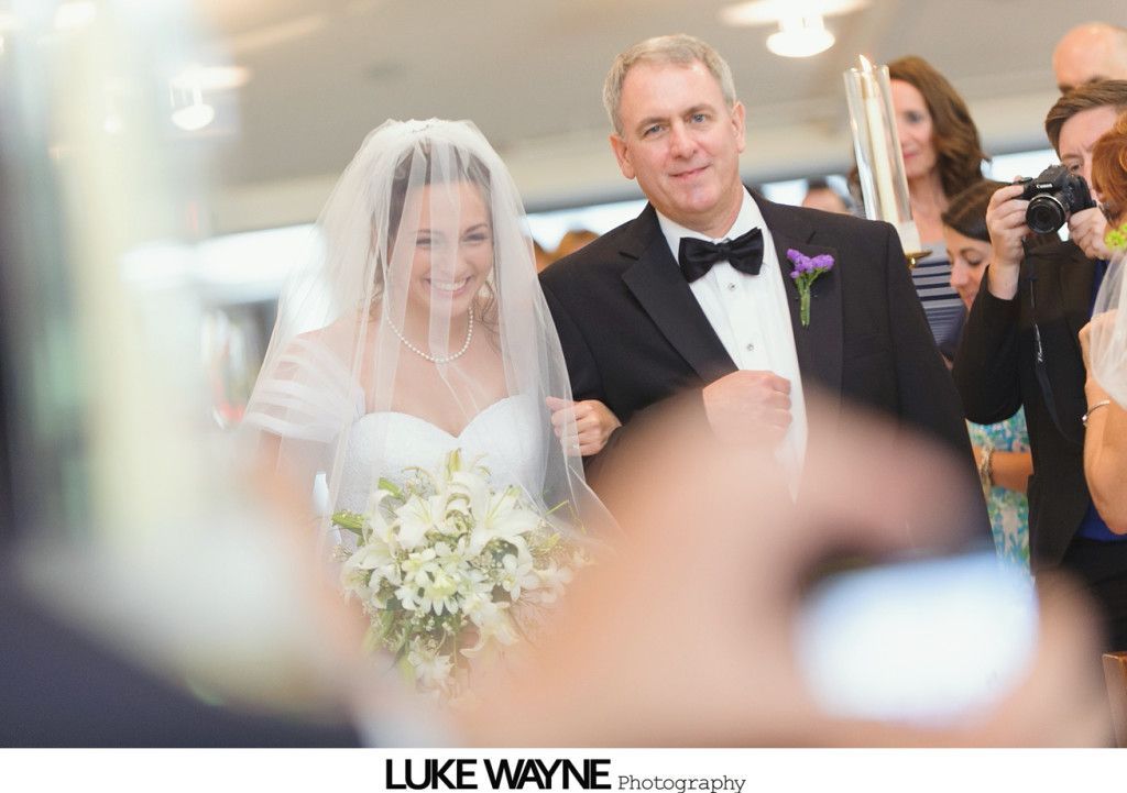 Bride in white dress and veil walks down the aisle with her father, holding a bouquet.