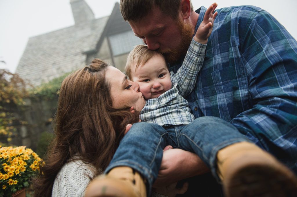 Parents kissing a smiling child outdoors near a stone building.