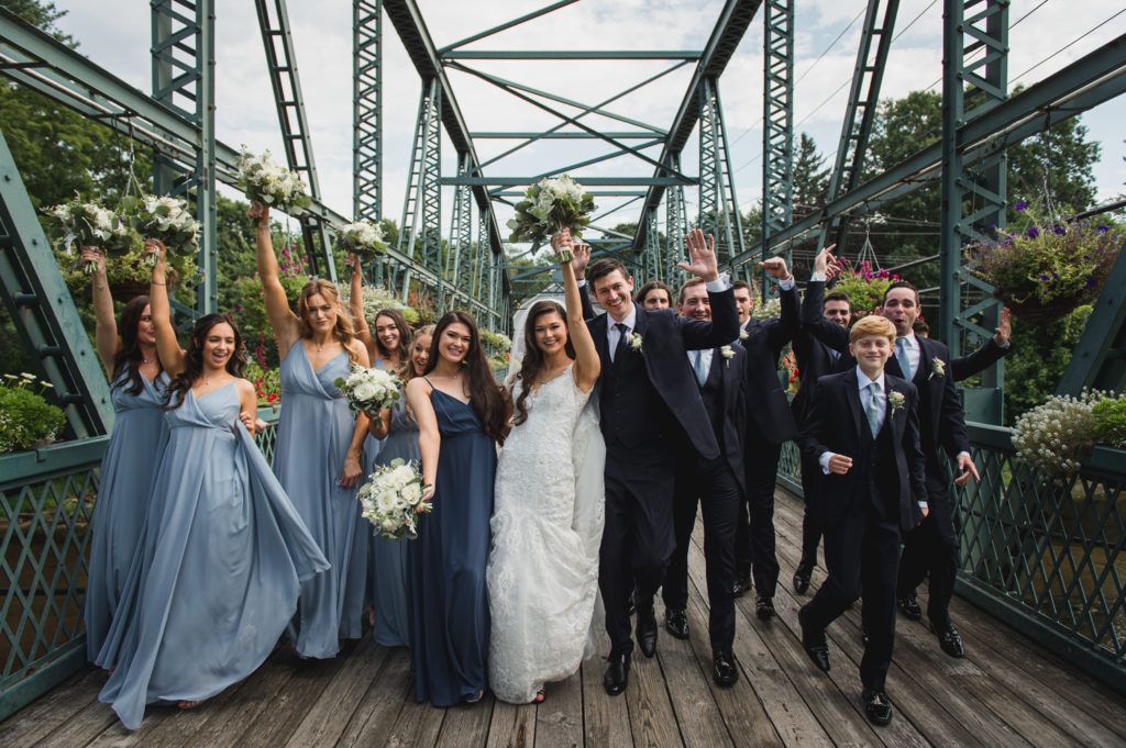 Wedding party on a green bridge, cheering with bouquets raised.  Bride and groom in center, bridesmaids in blue dresses, groomsmen in suits.