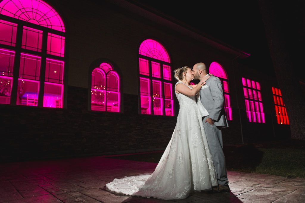 Couple kissing in front of a building with pink-lit arched windows at night.