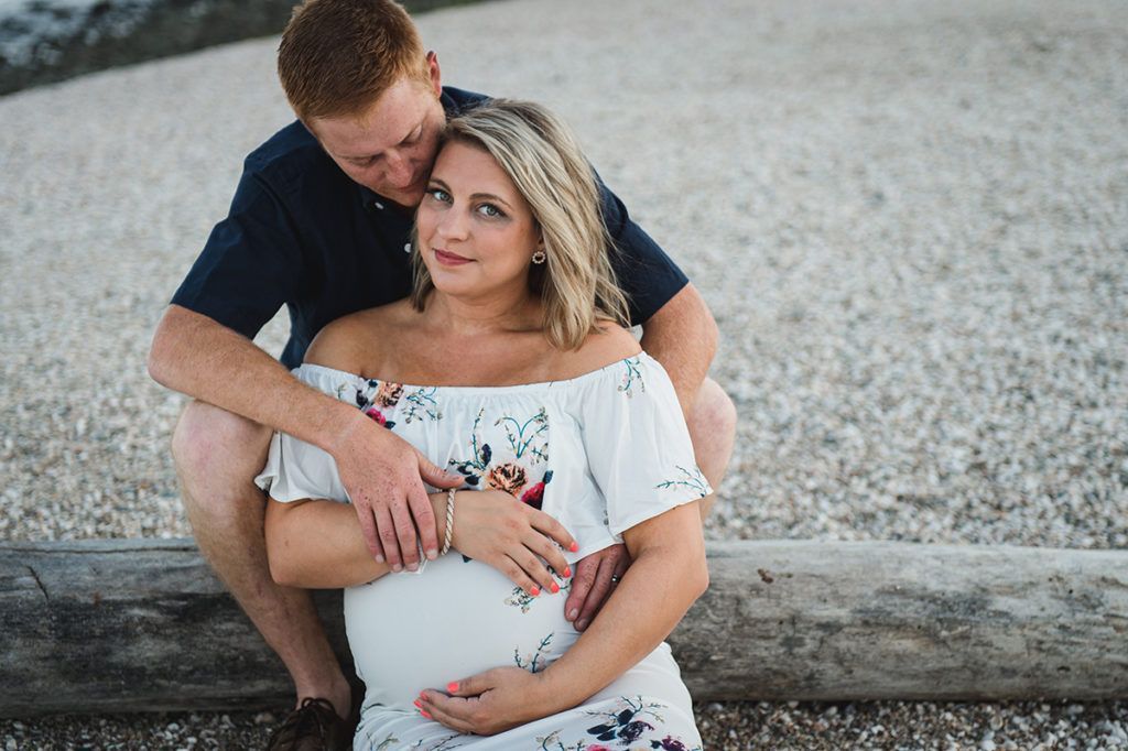 Man hugs pregnant woman; outdoors, sitting on log. He wears a navy shirt, she has on a floral dress.