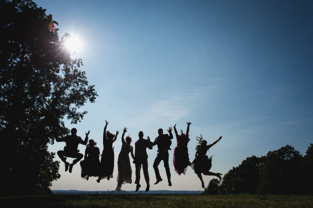Silhouetted group jumping on a grassy hill, arms raised against a bright sun and blue sky.