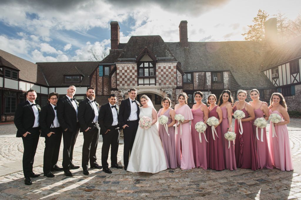 Wedding party poses in front of a Tudor-style building. Bride in white gown with bridesmaids in pink dresses, groomsmen in black tuxedos.