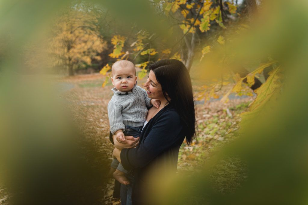 Woman holding a baby outdoors, both looking at each other, surrounded by fall foliage.