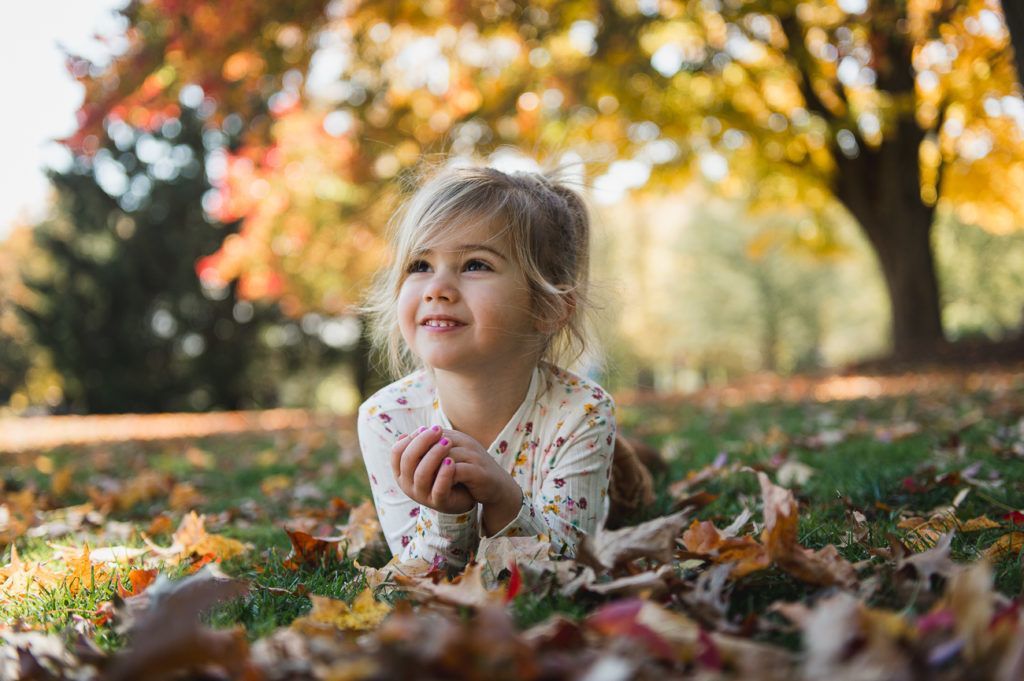 Young child lying in leaves, smiling, in front of a tree with fall foliage.