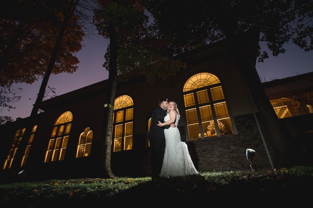 Couple kissing in front of a building with arched windows lit from within, at dusk.