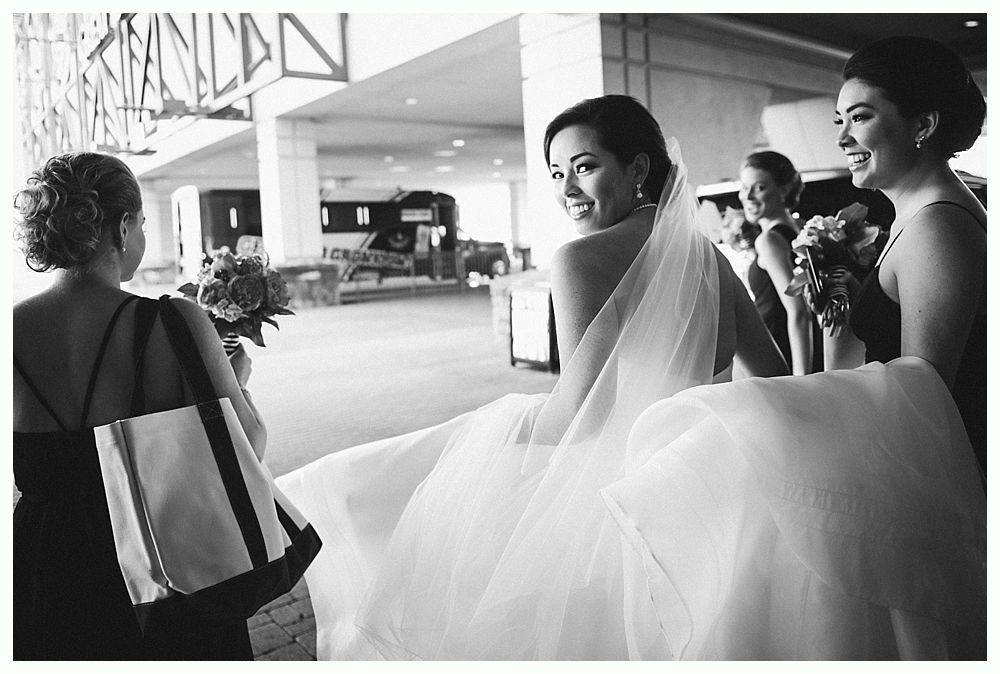 Bride in a white gown smiles, looking over her shoulder. Bridesmaids in dark dresses hold bouquets outside a building.