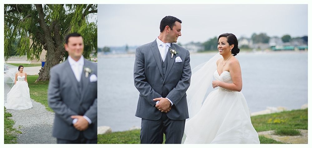 Wedding couple; groom in gray suit, bride in white dress, by water. Bride smiles, groom looks on.