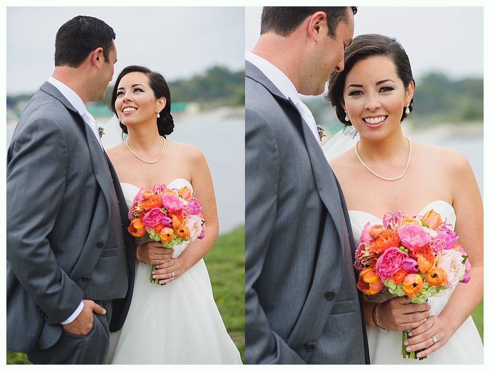 Bride and groom smiling, holding flowers. Gray suit, strapless white dress, lake background.