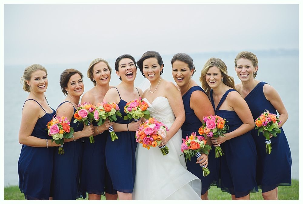 Bridesmaids in navy dresses laugh, holding colorful bouquets next to the bride in a white gown by the water.