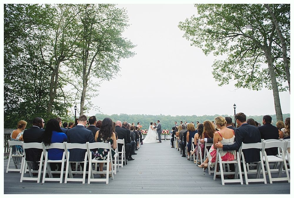 Wedding ceremony outdoors, guests seated on either side of a walkway leading to a couple at the altar. Trees frame the view.