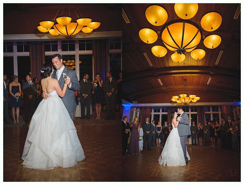 Bride and groom dance at reception under ornate chandeliers. Guests watch in a wood-paneled room.