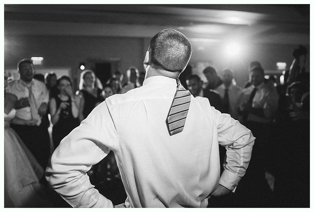 Man at a party with arms on hips, tie askew, looking up, with a crowd of people in the background.