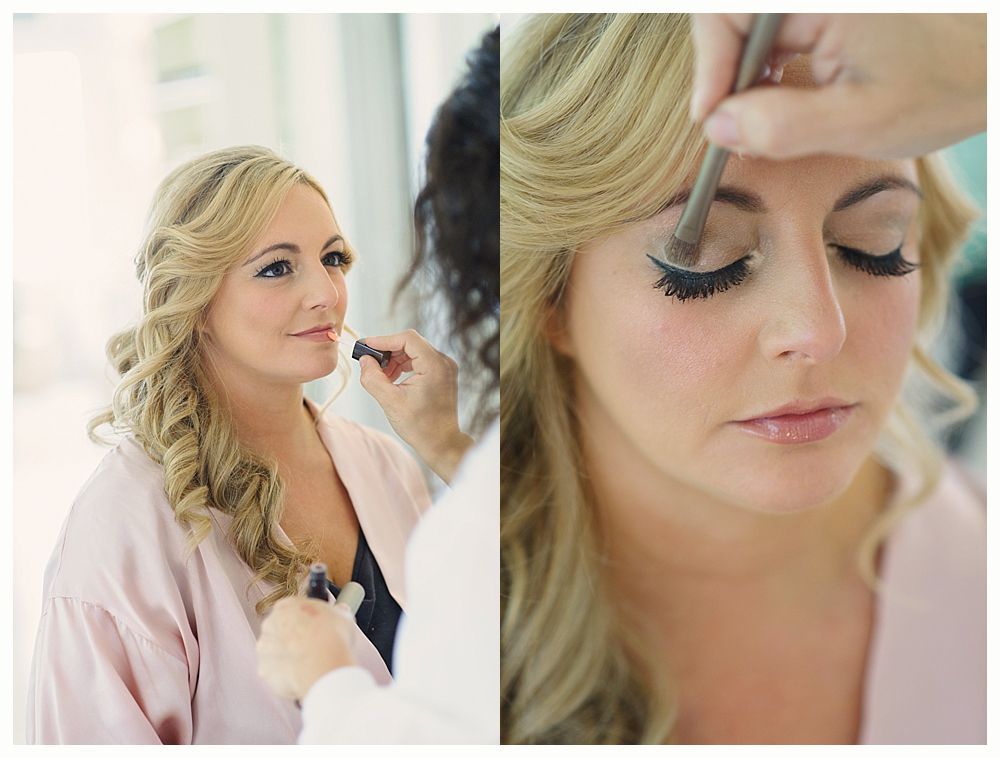Woman getting makeup done, wearing a pink robe. Makeup artist applies eye shadow. Blonde hair in curls.