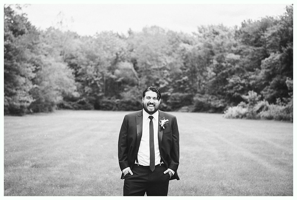 Man in a tuxedo smiles, standing in a field with trees in the background. Black and white photo.