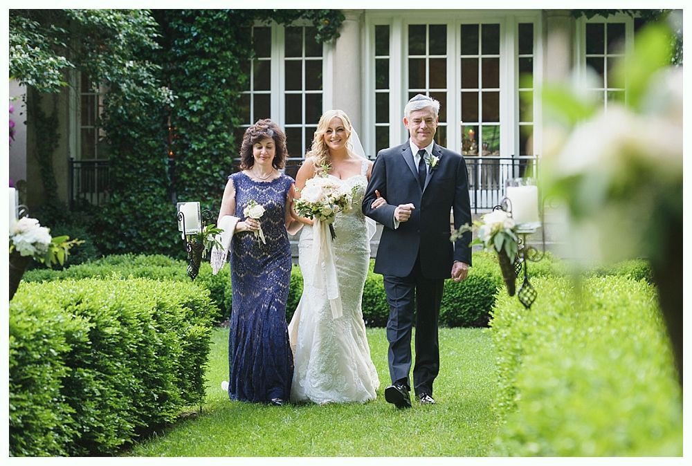 Bride walking down garden aisle with parents. She wears white lace gown, they're in formal attire. Green hedges and white flowers line the path.
