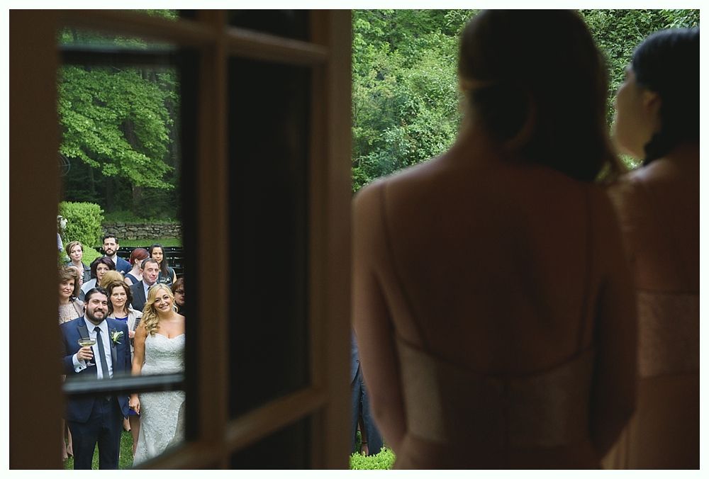 Bridesmaids watch a wedding ceremony through a window. The couple stands in a garden among guests.