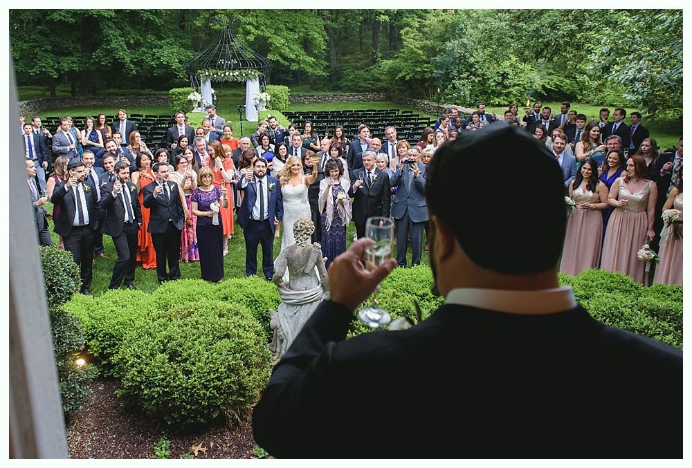 Person toasting, overlooking wedding guests gathered in a garden, greenery and trees in background.