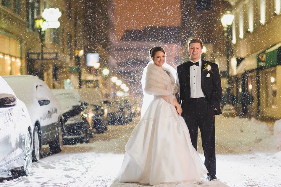 Bride and groom in formal wear pose in snowy street, surrounded by parked cars and buildings.