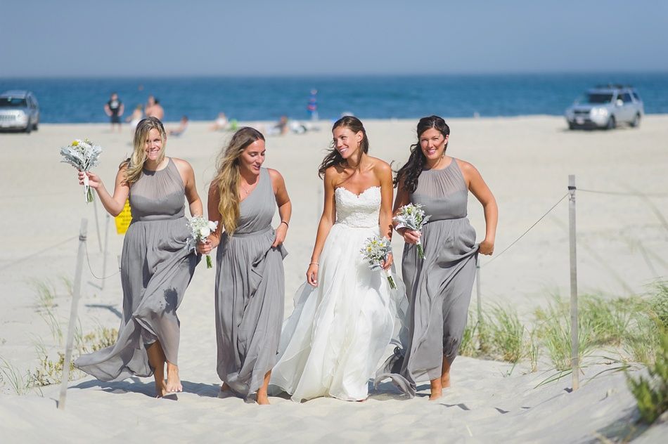 Bride and bridesmaids walk barefoot on a sandy beach. They wear gowns: white for the bride, gray for bridesmaids. Ocean in the background.