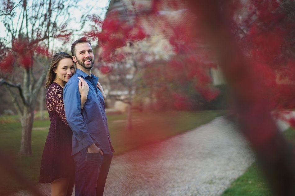 Couple embracing outdoors, framed by red blossoms. Man smiles, woman rests hand on his shoulder. Path and trees in background.