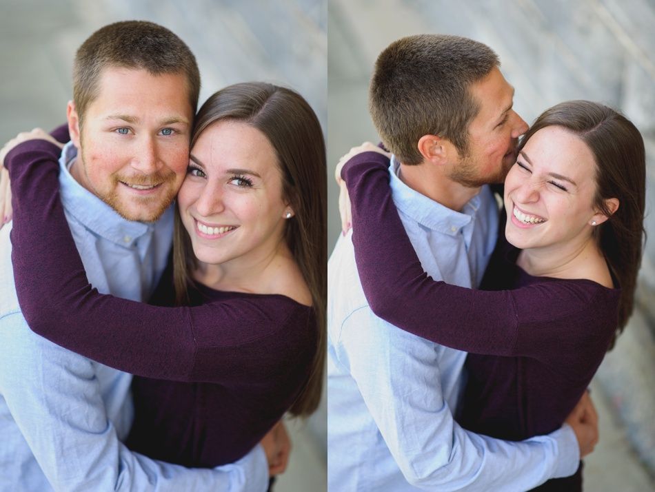 Couple embraces, smiling. Man kisses woman's cheek. Both wear long-sleeved shirts, posing near stairs.
