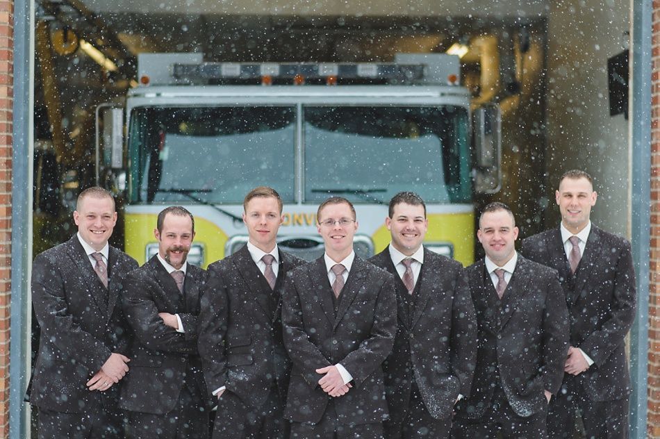 Group of men in suits in front of a fire truck in snowy conditions, inside a fire station.