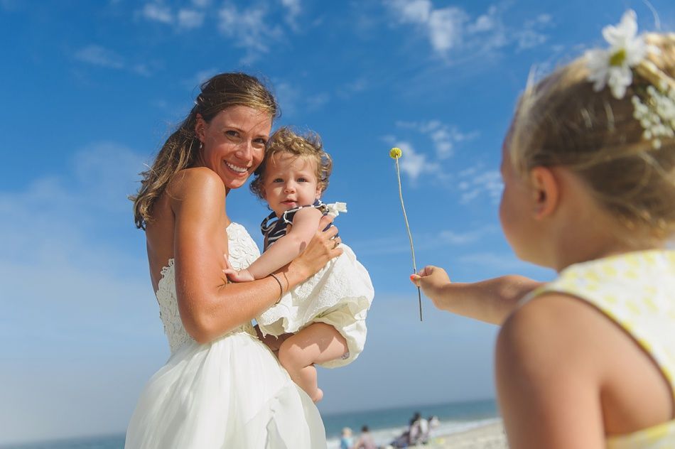 Bride holding baby on beach, looks at child holding flower. Blue sky, bright sunlight.
