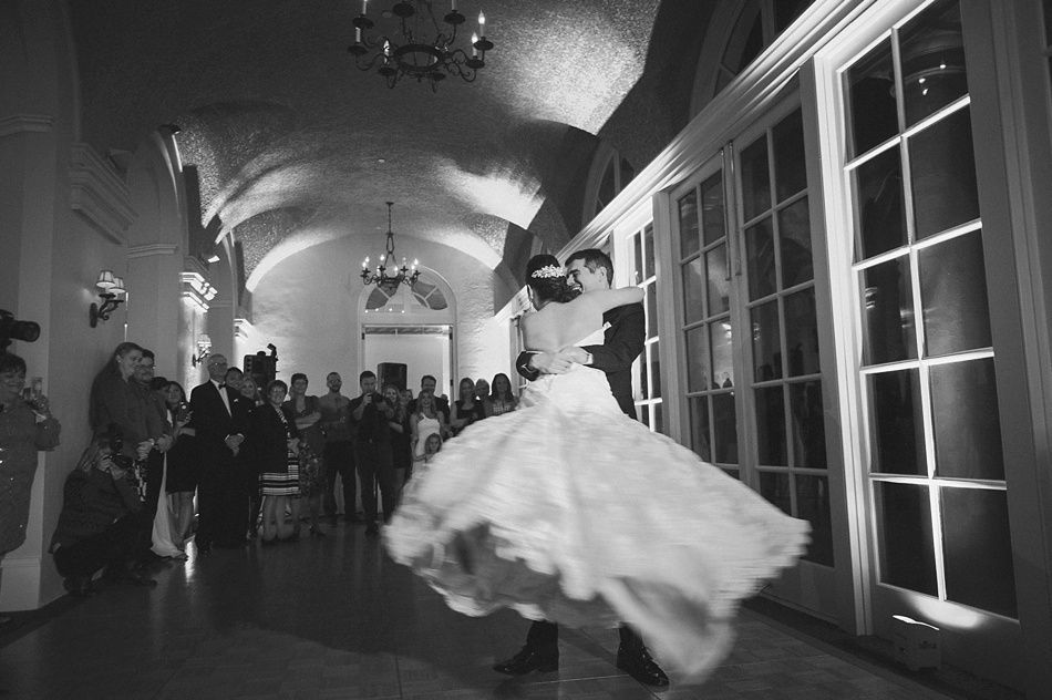 Bride and groom dance at wedding reception. Ballroom, bride's dress swirling, guests watch. Black and white.