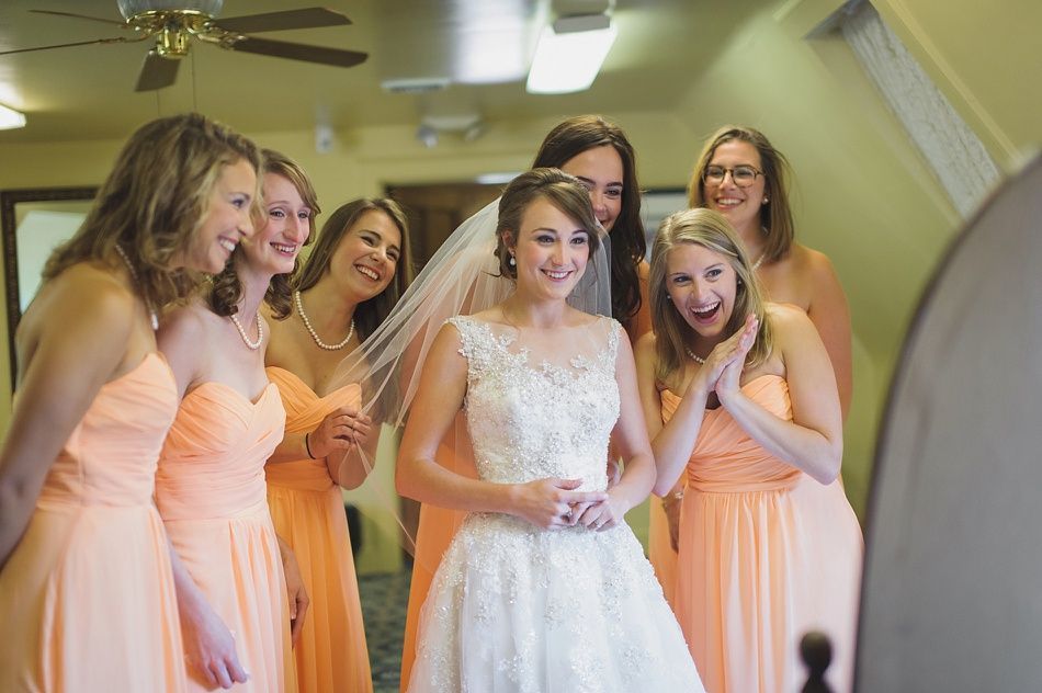 Bride in a white dress, veil, with bridesmaids in peach dresses smiling, looking at a mirror in a room.