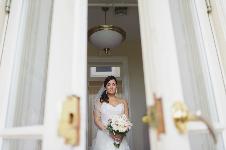 Bride in a white dress and veil holding flowers, standing between open doors, looking at camera.
