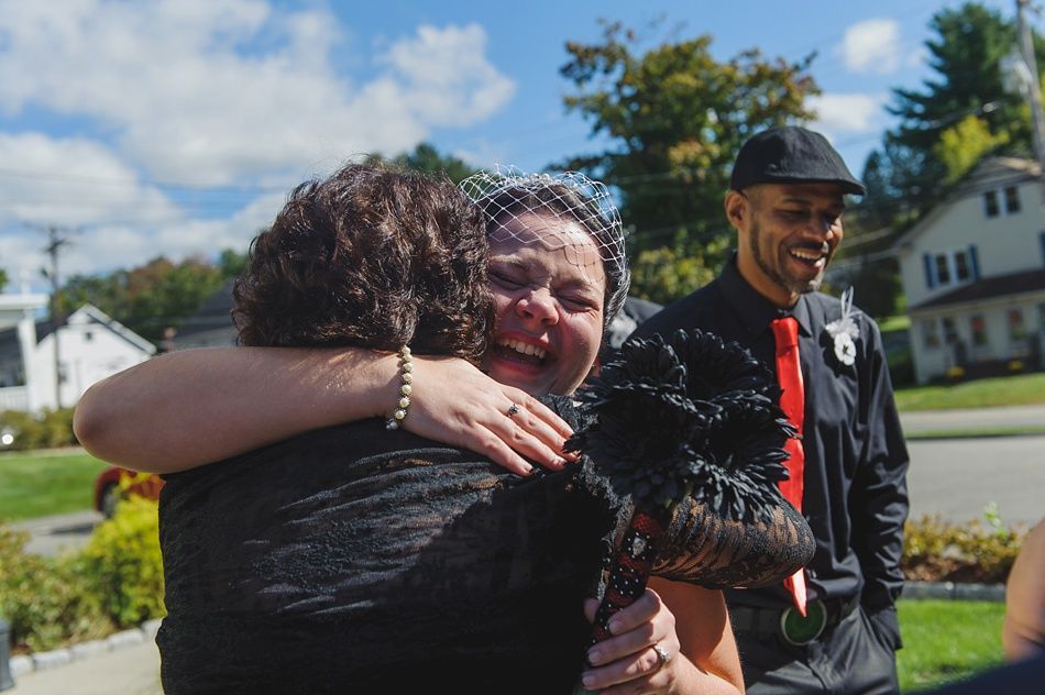 Woman in veil hugs another woman, both smiling; man in black suit smiles nearby. Outdoors.