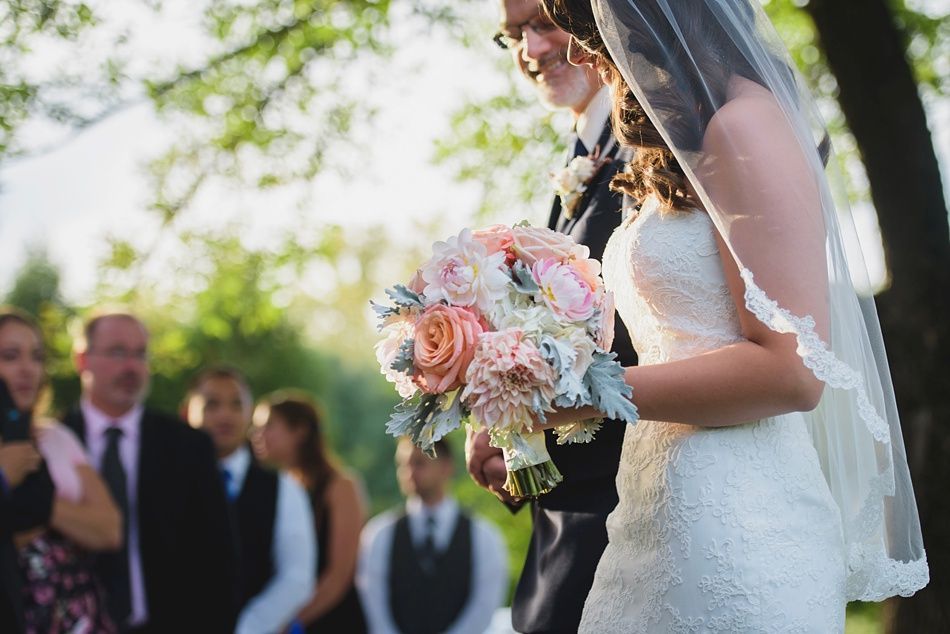 Bride walking down the aisle with a bouquet, father beside her, attendees watch in a sunlit outdoor setting.