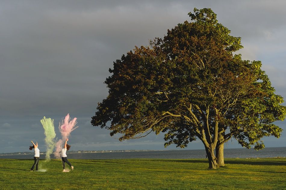 Two people throwing colored powder near a large tree on a grassy field, overcast sky.