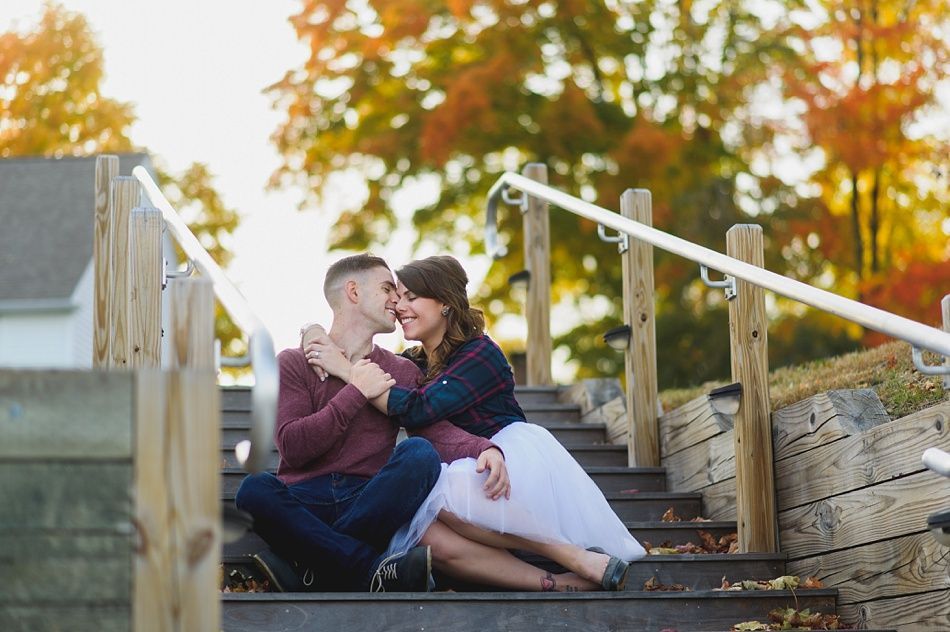 Couple kissing on outdoor steps with fall foliage. Man in maroon shirt, woman in plaid and white skirt.