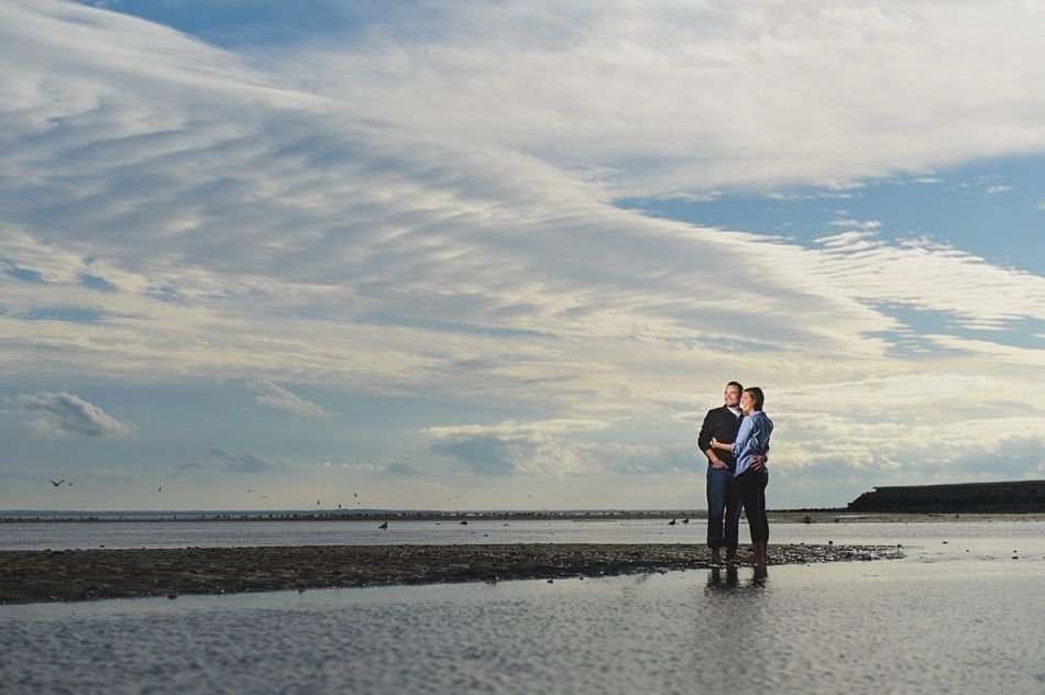 Couple standing in shallow water, looking up at cloudy sky over a beach.