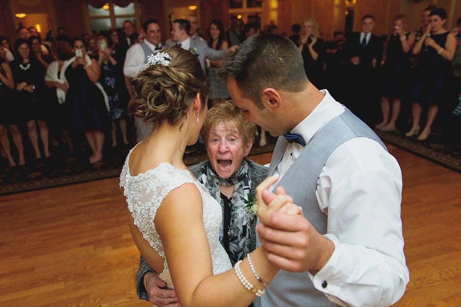 Bride and groom dance at reception with a surprised woman in the middle; guests watch.