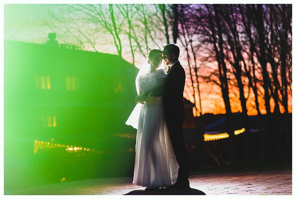Couple embracing at sunset, bride in white dress, groom in suit. Trees, house in background.