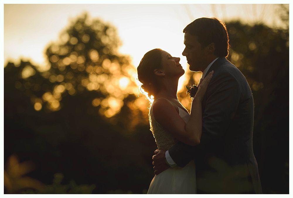 Couple embracing at sunset, bride in white dress, groom in suit. Golden light in background.