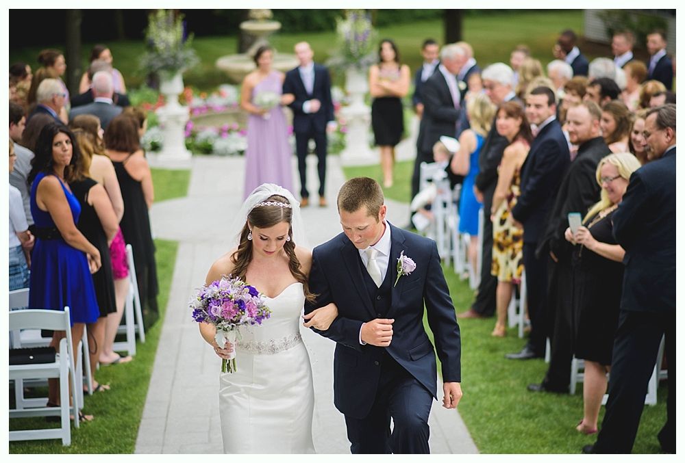 Bride and groom walk down aisle, wedding ceremony. Guests watch; venue outdoors with white chairs and green lawn.