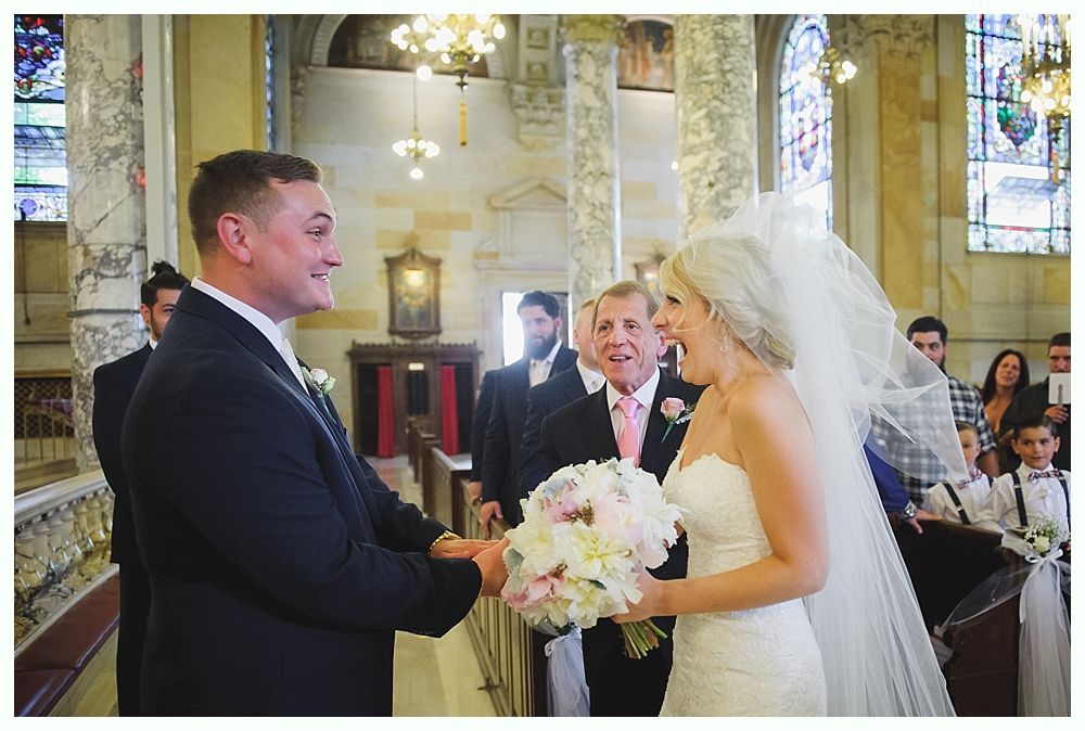 Bride and groom exchanging vows at altar, church setting. Bride holds flowers, groom smiles. Guests watch.