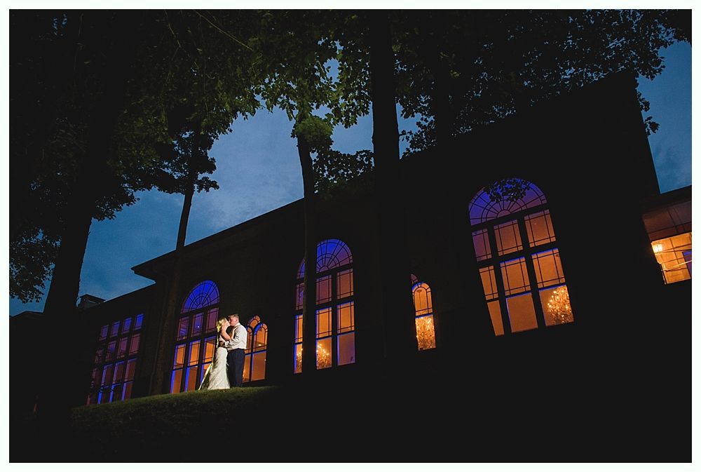 Couple embraces in front of a building with arched windows lit with blue and orange light at dusk.