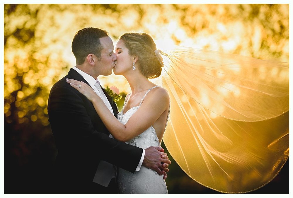 Couple kissing, bride's veil billowing in golden sunlight, groom's hand on her back, formal attire.