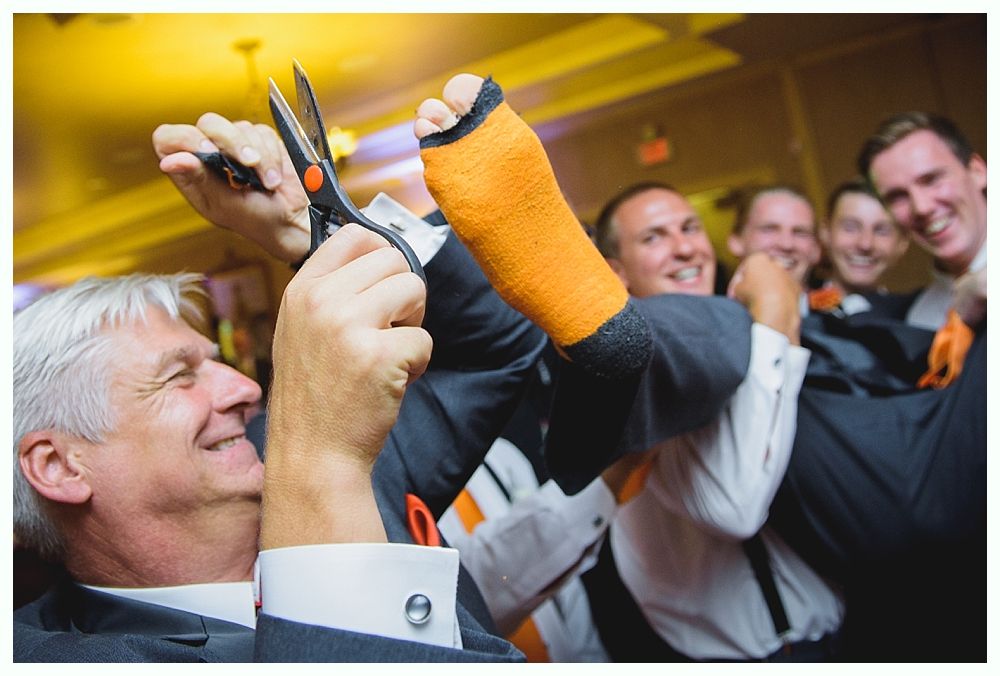 Man cutting cast off another man's leg with scissors. Group of men cheering in a brightly lit room. Orange cast.