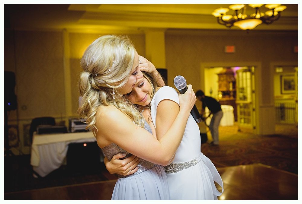 Two women embracing, one holding a microphone. Dimly lit room with a chandelier and a person in the background.