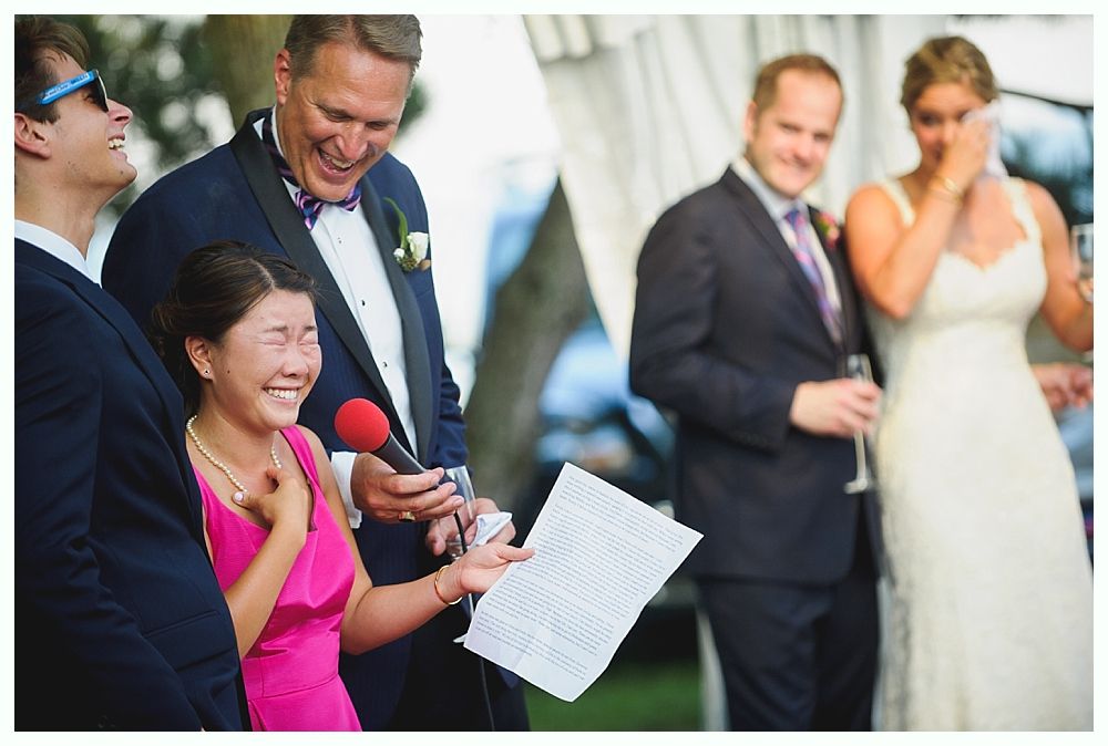 Wedding guests laughing and crying during a speech. Woman in pink dress holds microphone, others in suits. Bride in white dress.